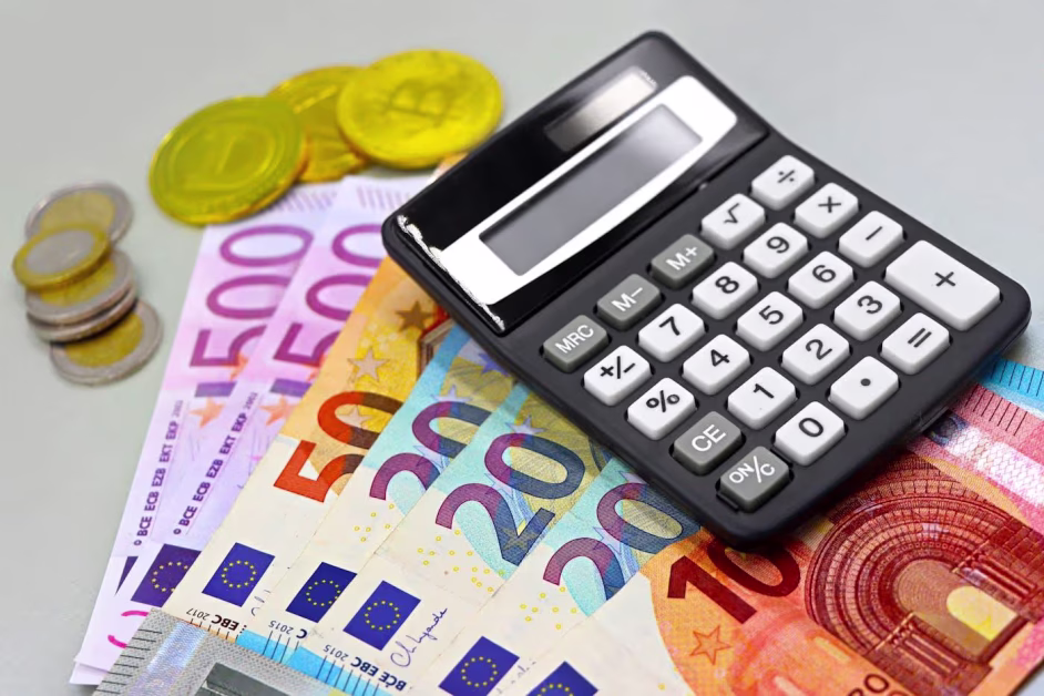 Une petite calculatrice noire posée sur une pile de billets de banque en euros (10, 20, 50, 500) à côté de pièces de monnaie.