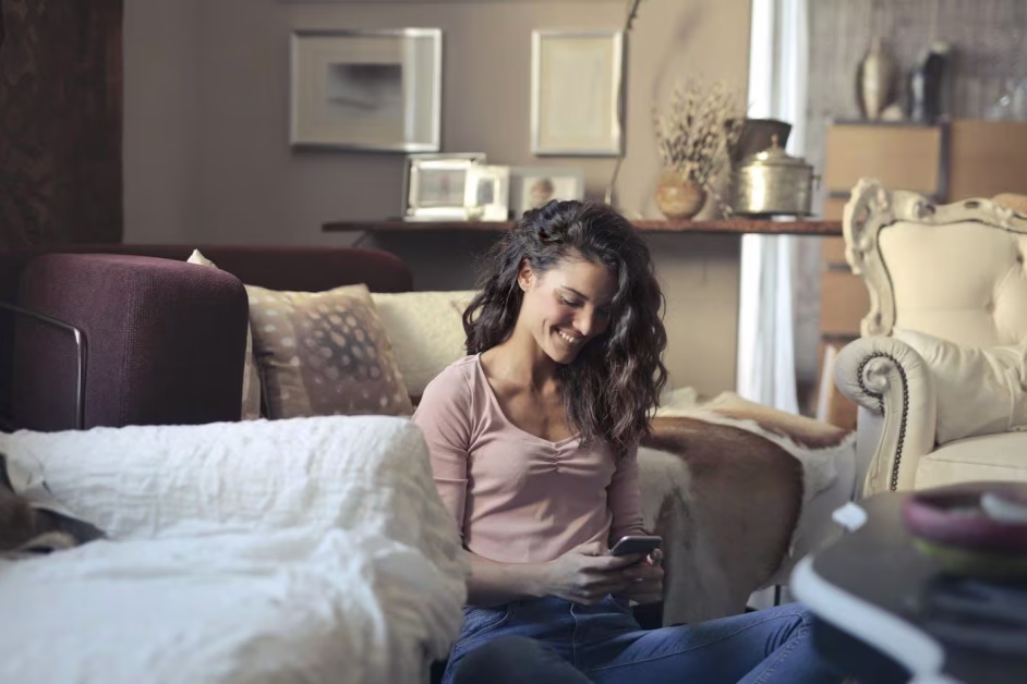 Une jeune femme souriante assise par terre dans son salon, en train de regarder l'écran de son smartphone.