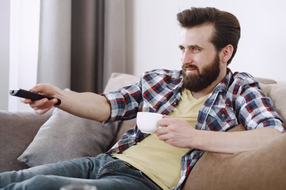 Homme barbu assis sur un canapé, pointant une télécommande d'une main et tenant une tasse blanche de l'autre.