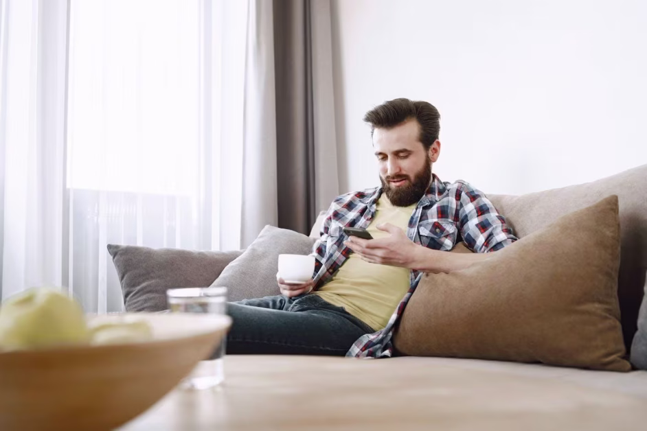 Homme barbu assis sur un canapé, consultant son smartphone tout en tenant une tasse blanche.