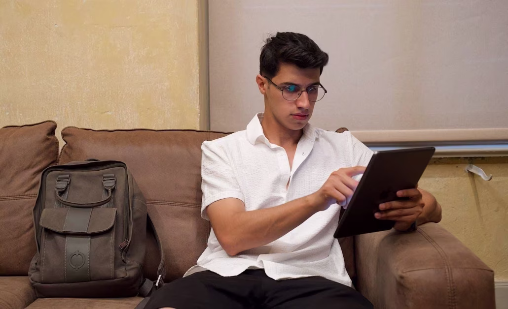 Jeune homme portant des lunettes assis sur un canapé en cuir marron et utilisant une tablette tactile.