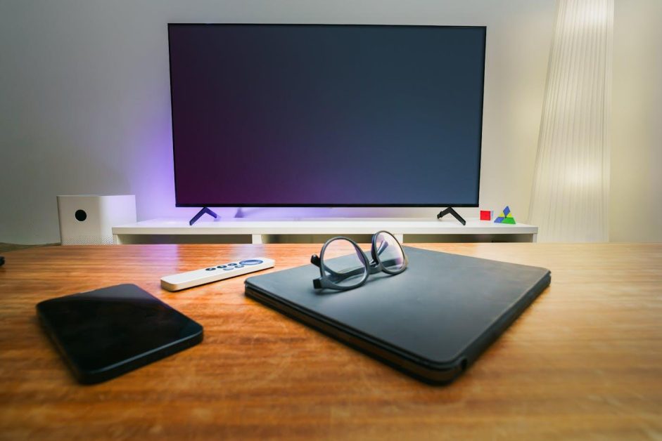 Une paire de lunettes de vue posée sur une pochette de tablette fermée, à côté d'un smartphone et d'une télécommande sur une table en bois face à une télévision éteinte.