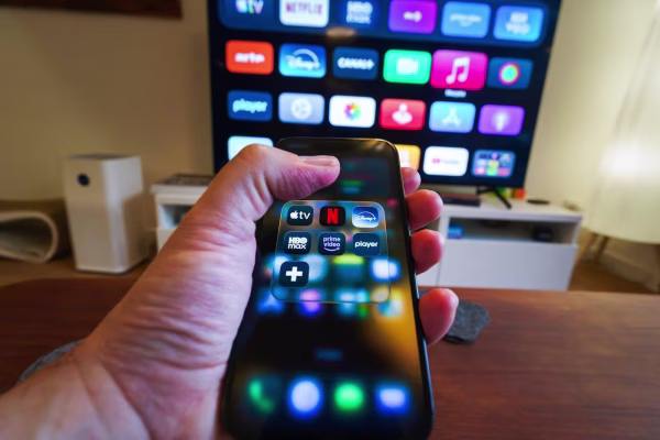Gros plan sur un smartphone affichant un dossier d'applications de streaming (Netflix, Prime Video, Disney+), avec un téléviseur connecté en arrière-plan.