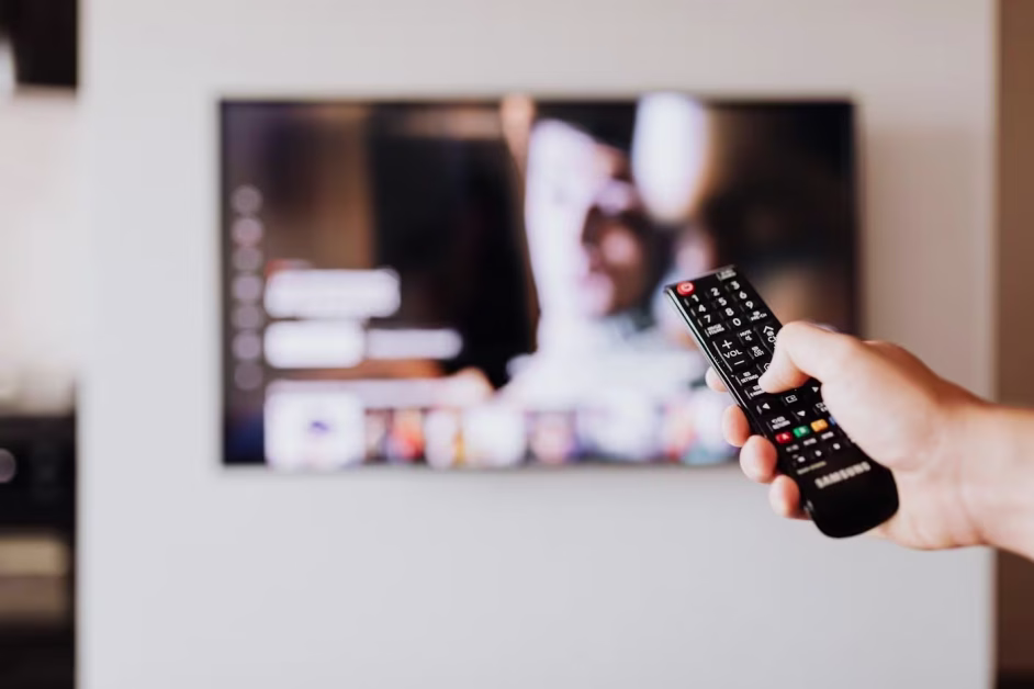 Une main au premier plan tenant une télécommande noire de marque Samsung pointée vers un écran de télévision flou.