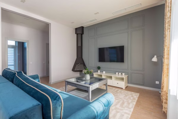 Grand salon lumineux équipé d'un canapé bleu en velours au premier plan, d'une table basse vitrée et d'un téléviseur fixé sur un mur gris mouluré.