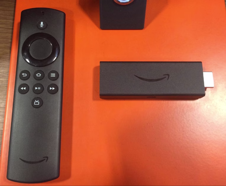 Télécommande noire et clé de streaming Amazon Fire TV Stick disposées sur un fond orange vif.